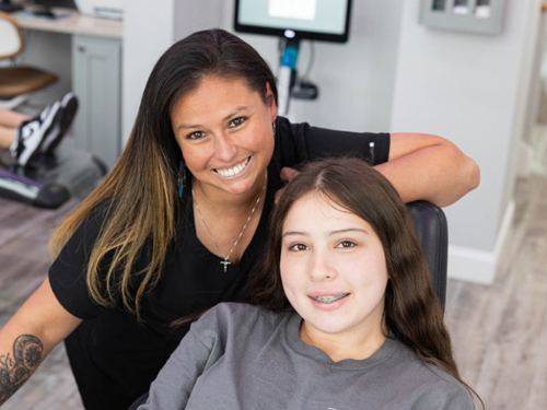Two people smiling at camera in orthodontic office setting with modern dental equipment visible.