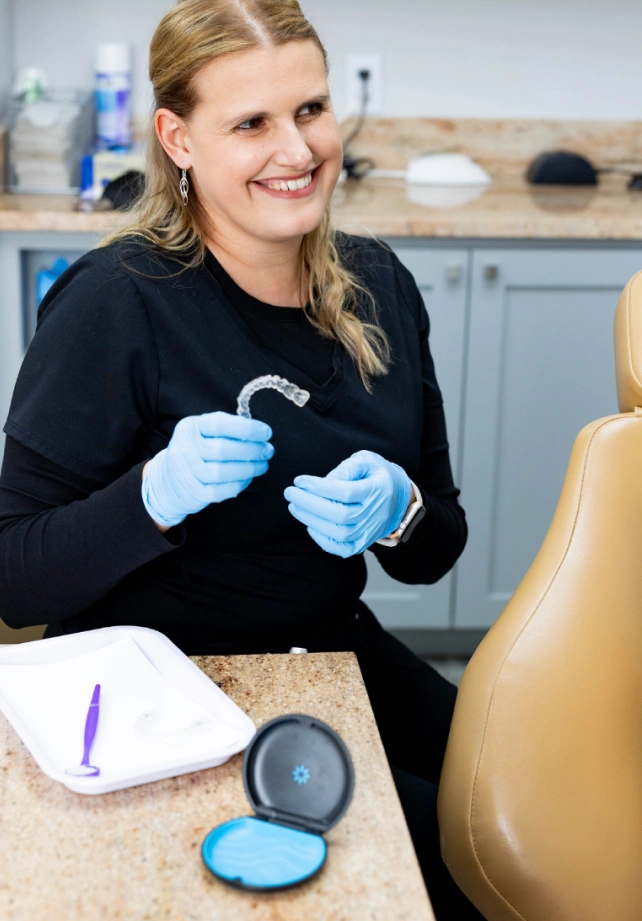 Orthodontist in clinical setting holding dental aligners and treatment materials at workstation.
